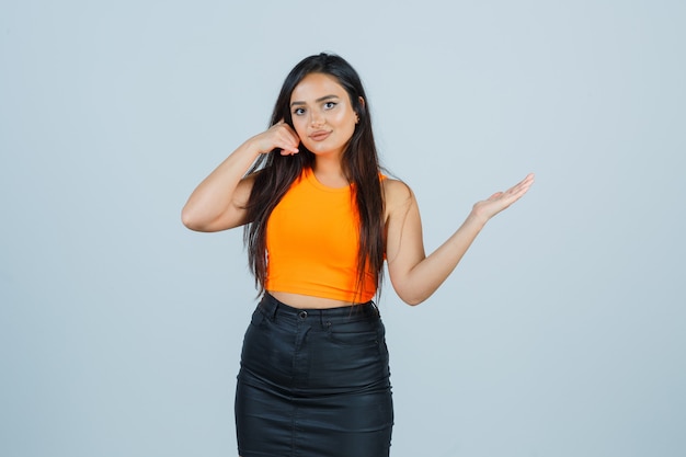 Señorita en camiseta, minifalda mostrando gesto de teléfono y mirando confiado, vista frontal.