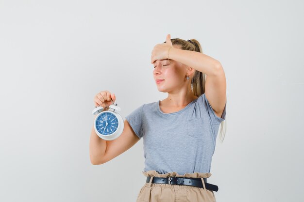 Señorita en camiseta y pantalón sosteniendo reloj despertador y mirando olvidadizo