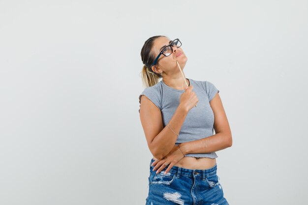 Señorita en camiseta, pantalones cortos sosteniendo un lápiz, mirando hacia arriba y mirando pensativo