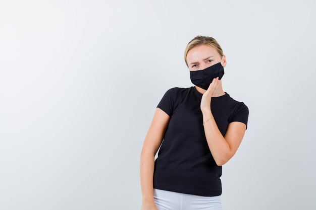 Señorita en camiseta, pantalones, máscara médica sosteniendo la mano en la máscara y mirando desconcertado