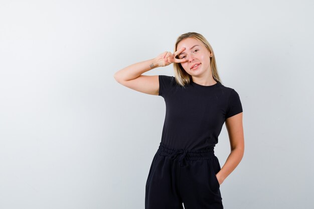 Señorita en camiseta, pantalones mostrando el signo de la victoria en el ojo y mirando alegre, vista frontal.