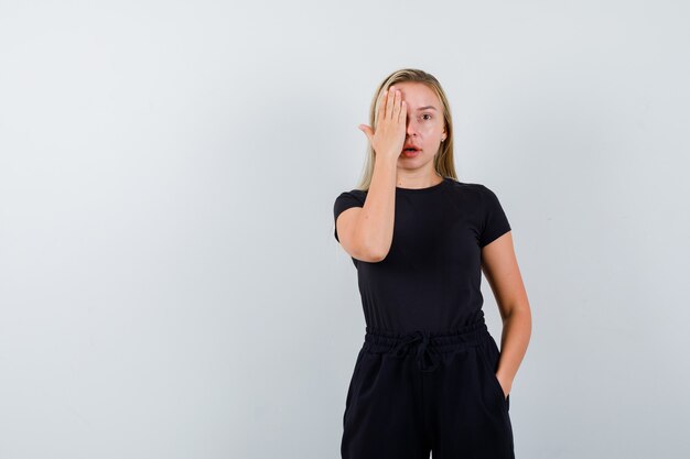 Señorita en camiseta, pantalones que cubren los ojos con la mano y mirando asombrado, vista frontal.