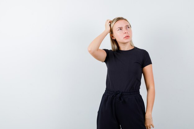 Señorita en camiseta, pantalones sosteniendo la mano en la cabeza y mirando pensativo, vista frontal.