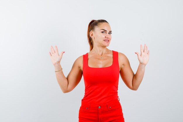 Señorita en camiseta roja, pantalón rojo mostrando las palmas de las manos, mirando a la cámara y mirando alegre, vista frontal.