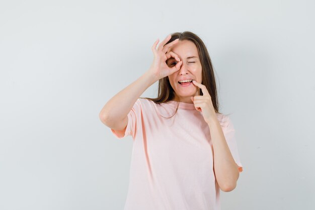 Señorita en camiseta rosa mostrando gesto ok en el ojo y mirando contento