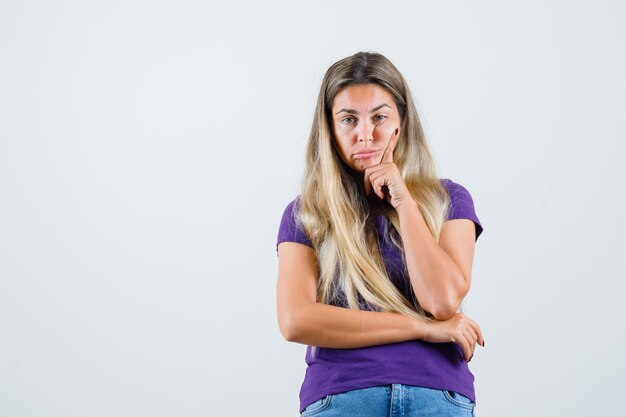 Señorita en camiseta violeta, jeans de pie en pose de pensamiento y mirando irresoluto, vista frontal.