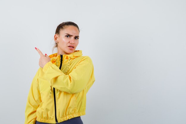 Señorita con chaqueta amarilla apuntando a la esquina superior izquierda y mirando insatisfecho, vista frontal.