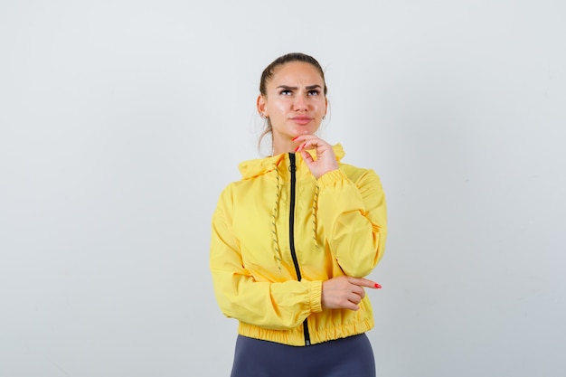 Señorita de chaqueta amarilla con la mano debajo de la barbilla y mirando pensativo, vista frontal.