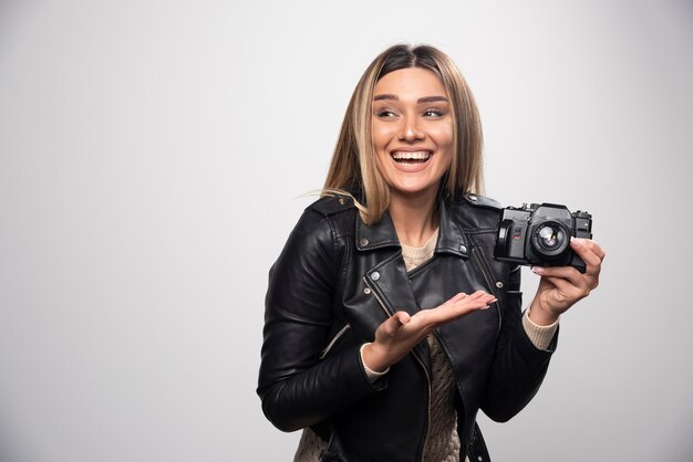 Señorita en chaqueta de cuero negro tomando fotos con cámara de manera positiva y sonriente.