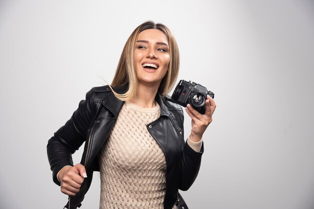 Señorita en chaqueta de cuero negro tomando fotos con cámara de manera positiva y sonriente.