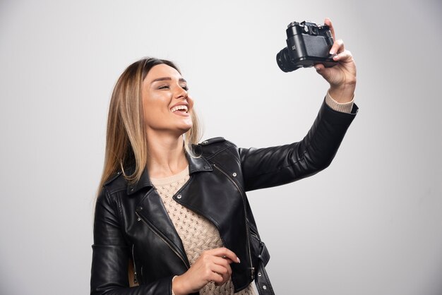 Señorita en chaqueta de cuero negro tomando fotos con cámara de manera positiva y sonriente.