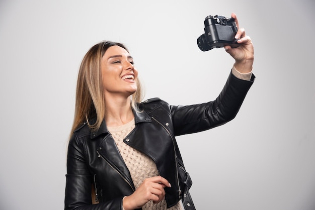 Señorita en chaqueta de cuero negro tomando fotos con cámara de manera positiva y sonriente.
