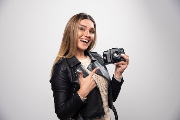 Señorita en chaqueta de cuero negro tomando fotos con cámara de manera positiva y sonriente