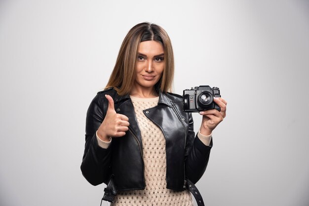 Señorita en chaqueta de cuero negro tomando fotos con cámara de manera positiva y sonriente