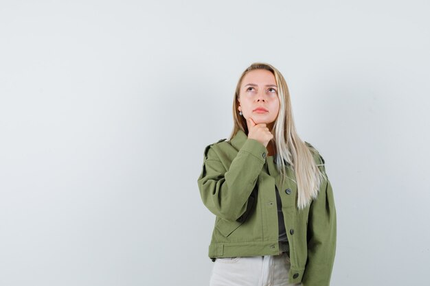 Señorita con chaqueta, pantalones apoyando la barbilla en la mano y mirando pensativa, vista frontal.