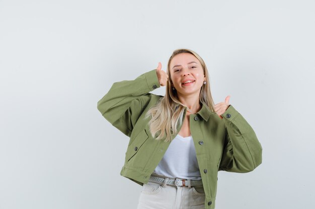 Señorita con chaqueta, pantalones mostrando el doble pulgar hacia arriba y mirando alegre, vista frontal.