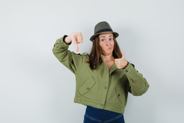 Señorita en chaqueta pantalones sombrero mostrando los pulgares hacia arriba y hacia abajo y mirando vacilante