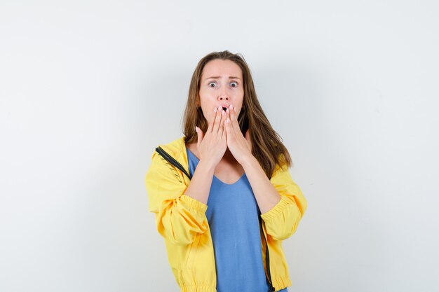 Señorita cogidos de la mano en la boca en camiseta, chaqueta y mirando consternado. vista frontal.