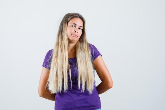 Foto gratuita señorita cogidos de la mano detrás de la espalda en camiseta violeta y mirando confiado, vista frontal.