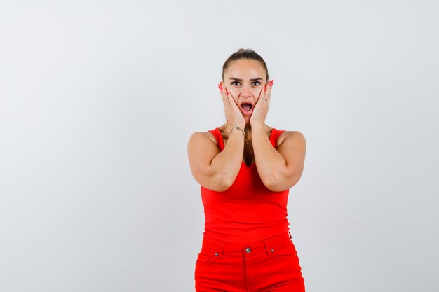 Señorita cogidos de la mano en las mejillas en camiseta roja, pantalón rojo y mirando perplejo, vista frontal.