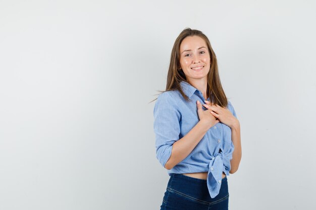 Señorita cogidos de la mano en el pecho con camisa azul, pantalones y mirando alegre