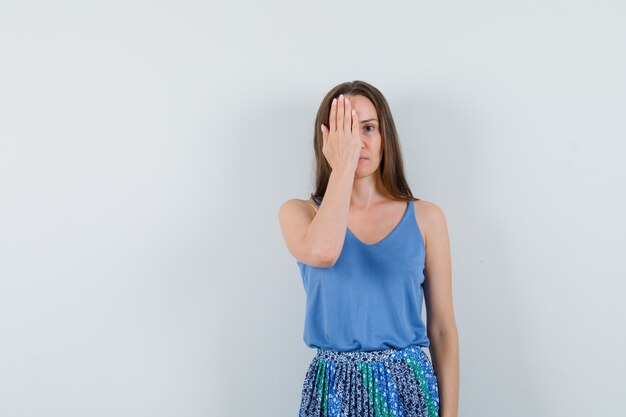 Señorita cubriendo su media cara con la mano en blusa azul, falda y mirando enfocado. vista frontal. espacio para texto