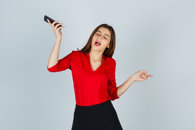 Señorita disfrutando mientras sostiene el teléfono móvil en blusa roja, falda y mirando divertido