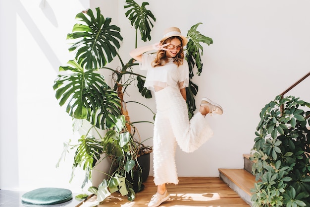 Señorita divertida en pantalones de moda de pie sobre una pierna y mostrando el signo de la paz con una sonrisa