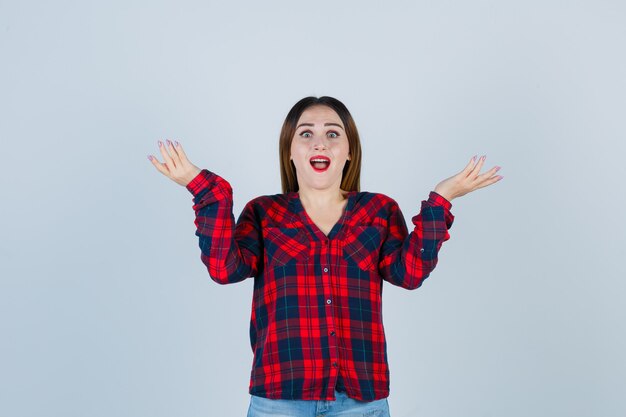 Señorita encogiéndose de hombros, abriendo la boca con camisa a cuadros y mirando confundida, vista frontal.