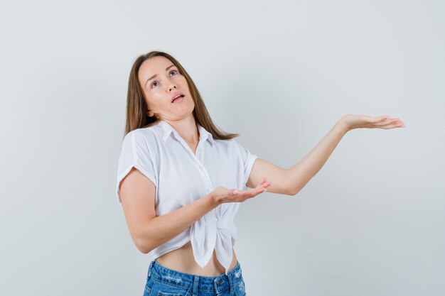 Señorita extendiendo sus palmas abiertas a un lado en blusa blanca y mirando disgustada. vista frontal.