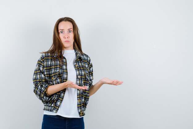 Señorita fingiendo mostrar algo en camiseta, chaqueta, jeans y mirando perplejo, vista frontal.