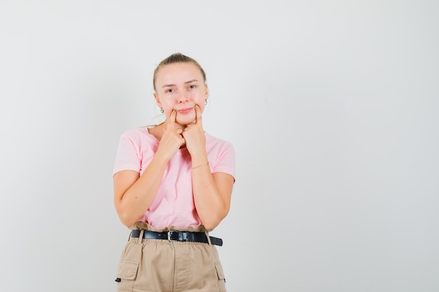 Señorita forzando una sonrisa en la cara en camiseta, pantalones y mirando descontento, vista frontal.