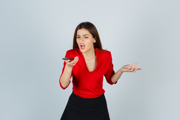 Señorita grabando mensaje de voz en el teléfono móvil en blusa roja, falda y con enojo