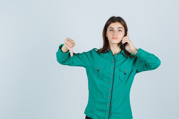 Señorita hablando por teléfono móvil, mostrando el pulgar hacia abajo en camisa verde y mirando disgustado, vista frontal.