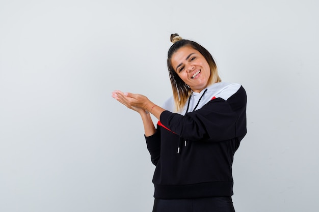 Señorita haciendo gesto de bienvenida en suéter con capucha y mirando feliz