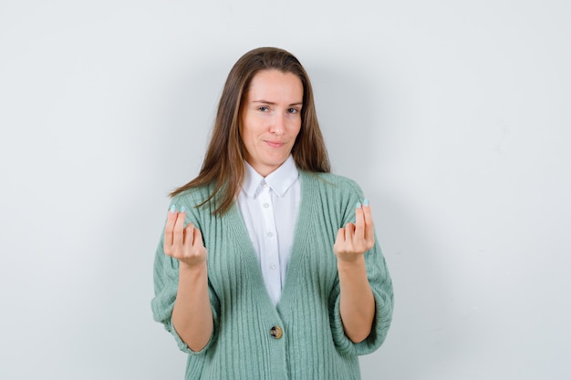 Señorita haciendo gesto de dinero en camisa, chaqueta de punto y mirando complacido. vista frontal.