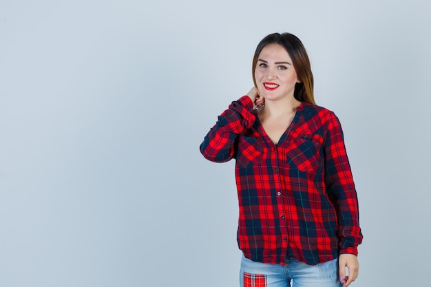 Señorita con la mano en el cuello en camisa a cuadros y mirando avergonzado, vista frontal.