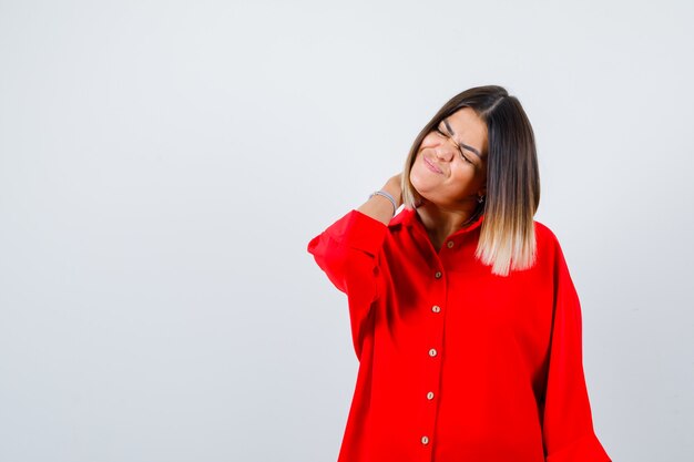 Señorita con la mano detrás del cuello en camisa roja de gran tamaño y luciendo dolorosa. vista frontal.