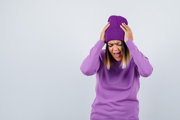 Señorita con las manos en la cabeza en suéter morado, gorro y mirando molesto. vista frontal.