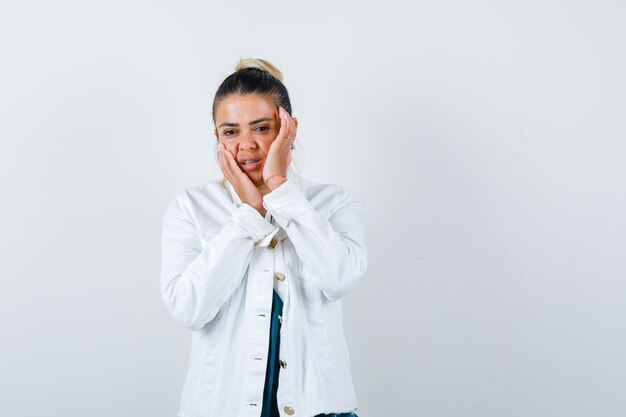 Señorita con las manos en las mejillas en camisa, chaqueta blanca y aspecto atractivo. vista frontal.