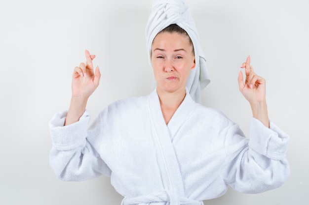 Señorita manteniendo los dedos cruzados en bata de baño blanca, toalla y mirando insatisfecho, vista frontal.