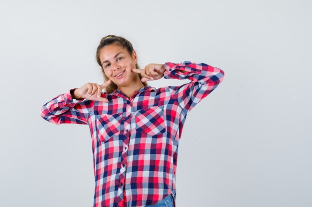 Señorita manteniendo los dedos en las mejillas en camisa a cuadros y mirando alegre. vista frontal.