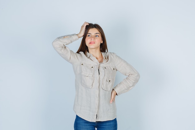 Señorita manteniendo la mano en la cabeza, mirando a un lado en casual, jeans y luciendo confundida. vista frontal.