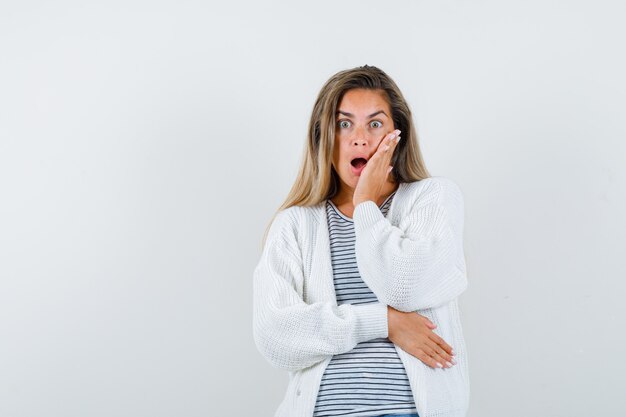 Señorita manteniendo la mano en la mejilla en camiseta, chaqueta y mirando sorprendido, vista frontal.