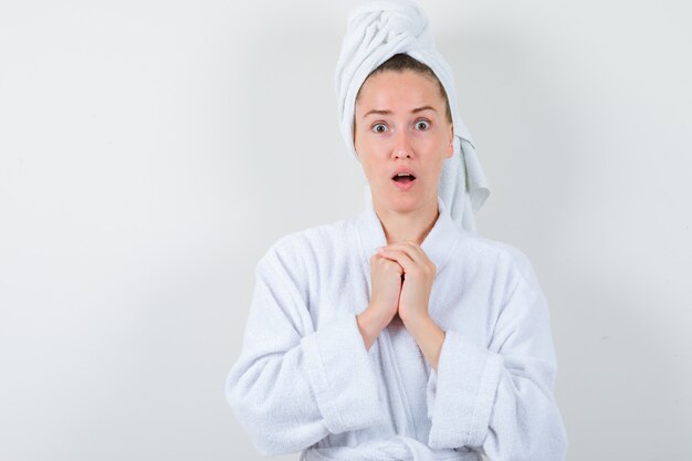 Señorita manteniendo las manos en el pecho en bata de baño blanca, toalla y mirando asombrado, vista frontal.