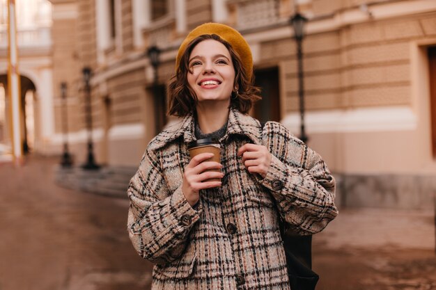 Señorita con maquillaje desnudo está sonriendo sinceramente contra la pared de la ciudad