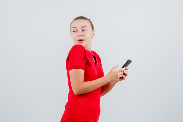 Señorita mirando hacia atrás mientras sostiene el teléfono móvil en camiseta roja y parece curioso.