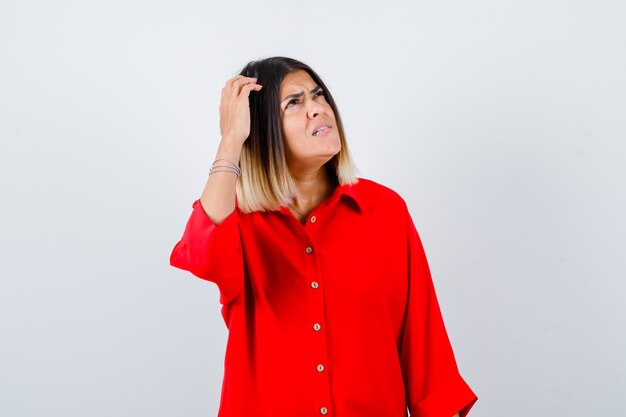 Señorita mirando a un lado mientras se rasca la cabeza con una camisa roja de gran tamaño y mira pensativa, vista frontal.