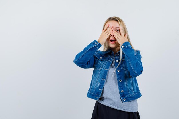 Señorita mirando a través de los dedos con ojos en camiseta, chaqueta vaquera, falda y luciendo linda