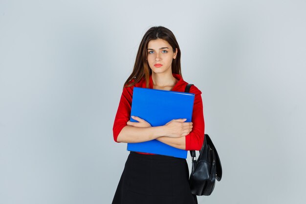 Señorita con mochila en blusa roja, falda sosteniendo una carpeta y mirando seria